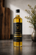A bottle of canola cooking oil with a black label and gold lettering, standing on a dark countertop in a kitchen setting with a wooden cutting board and a vase of flowers in the background.