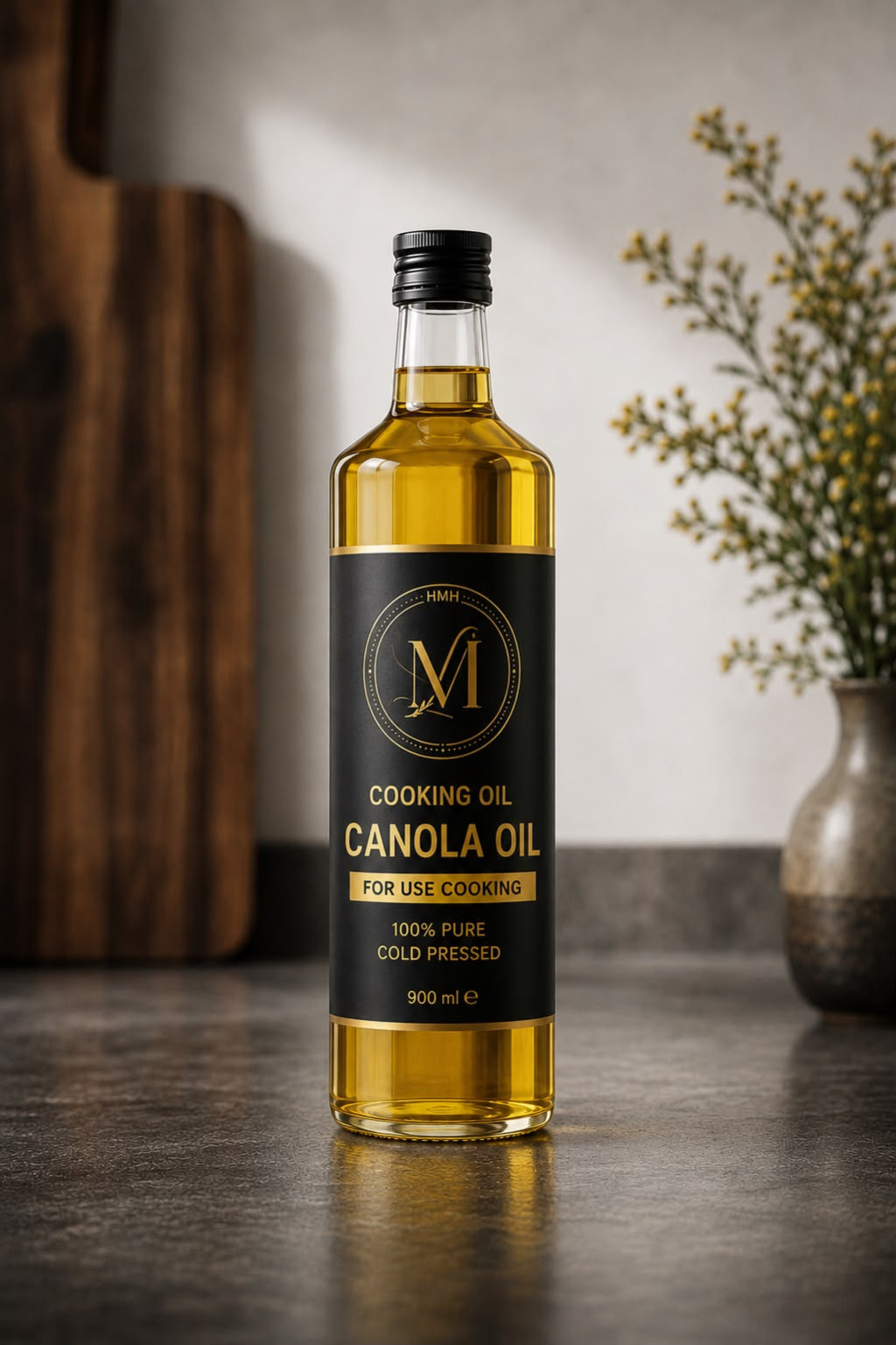 A bottle of canola cooking oil with a black label and gold lettering, standing on a dark countertop in a kitchen setting with a wooden cutting board and a vase of flowers in the background.
